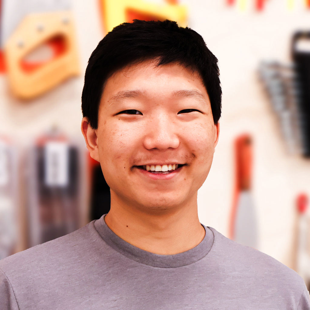 Smiling person standing in a workshop with various tools hanging on the wall in the background.